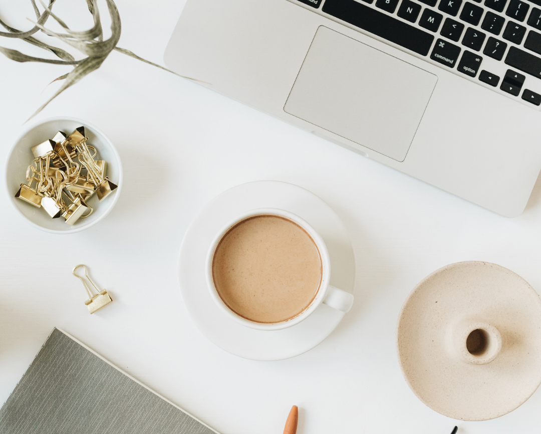 A cup of coffee sits on a saucer beside a laptop keyboard, a bowl of gold binder clips, an open notebook, and a plant on a white desk.