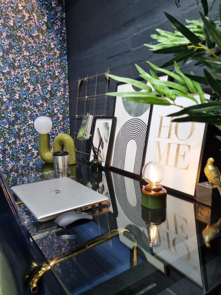 Laptop, mouse on a glass desk with decorative items and framed artwork, including one reading "HOME," against a dark wall with colorful floral wallpaper and green plant nearby.