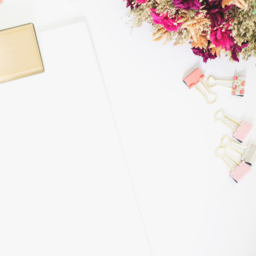 Golden binder, pink and floral binder clips scattered on white surface beside vibrant pink and beige dried flowers.