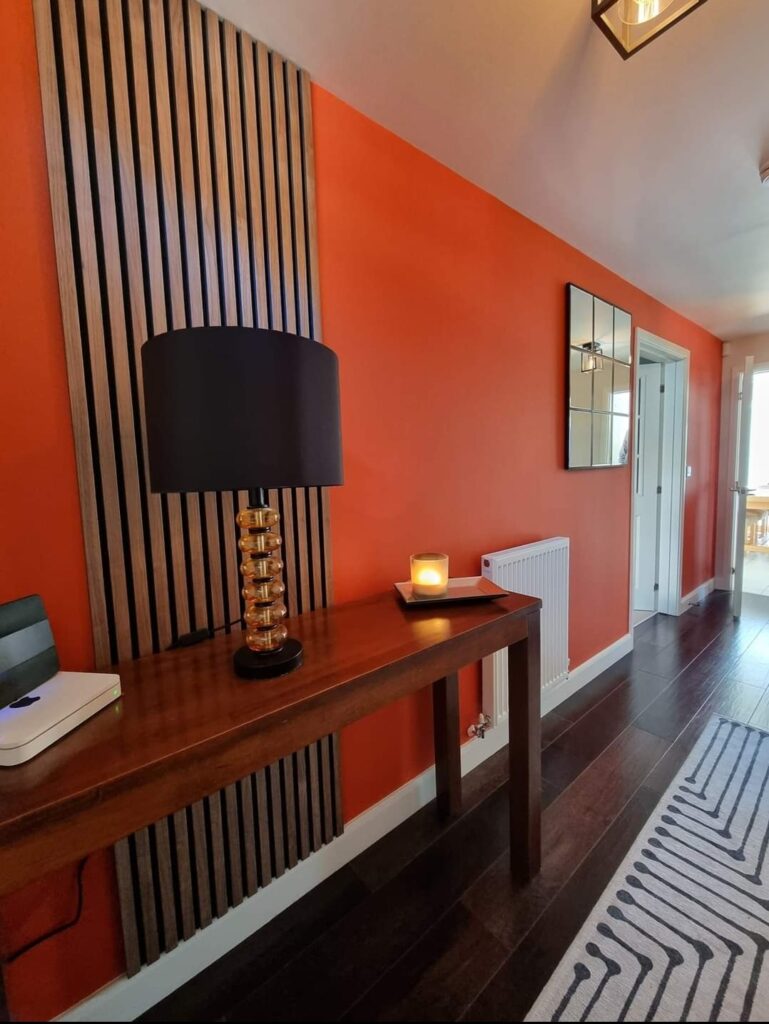 A wooden console table with a black lamp and a lit candle sits against an orange wall. Adjacent hallway features a mirror, a radiator, and a striped rug.