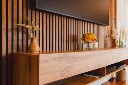 A wooden cabinet features vases with flowers and a candle, set against a wall with vertical wooden slats; a television is mounted above, and a remote control lies on the shelf.